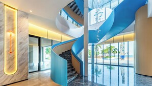 A modern interior with a curved staircase, marble walls, and large windows offering views of palm trees. - Holiday Inn Express Zhanjiang Coast by IHG (Zhanjiang)