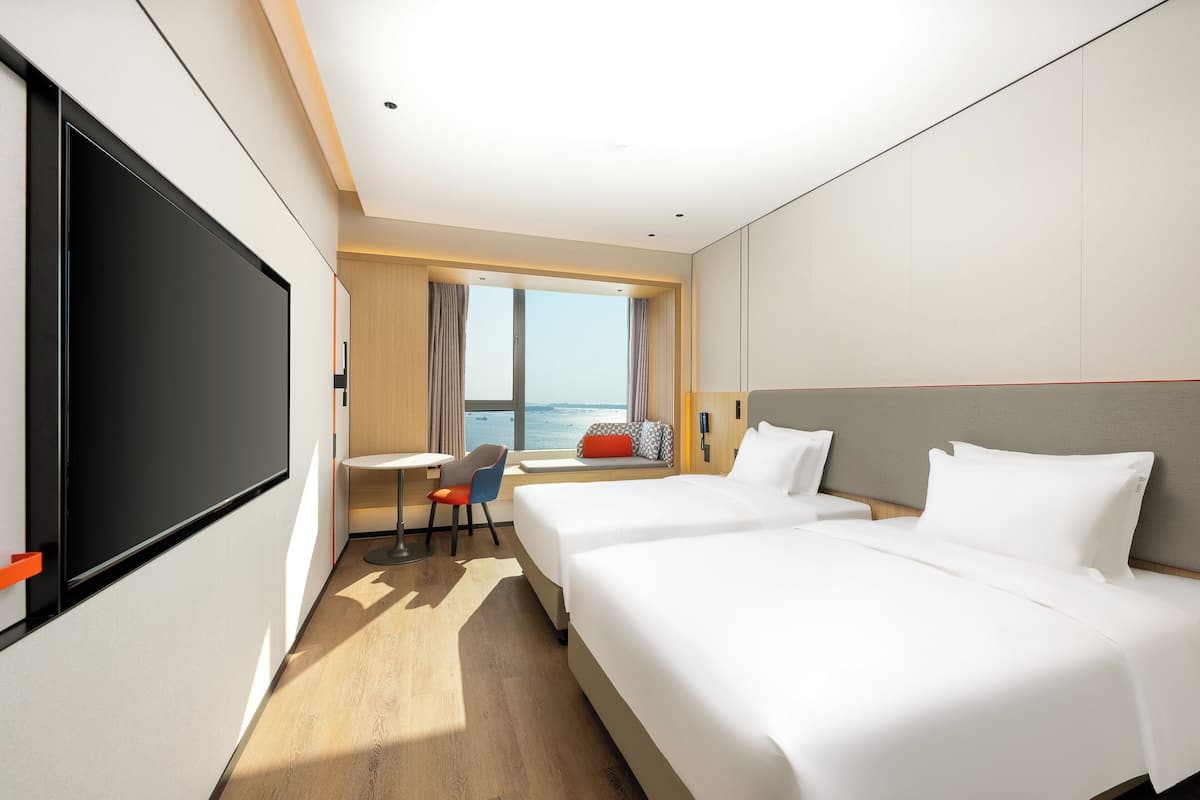Habitación de hotel con dos camas, un escritorio, una silla, televisión y ventana con vista al mar.