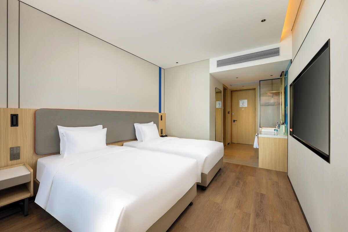 A hotel room with two beds, a desk, a bathroom with a mirror, and a TV.