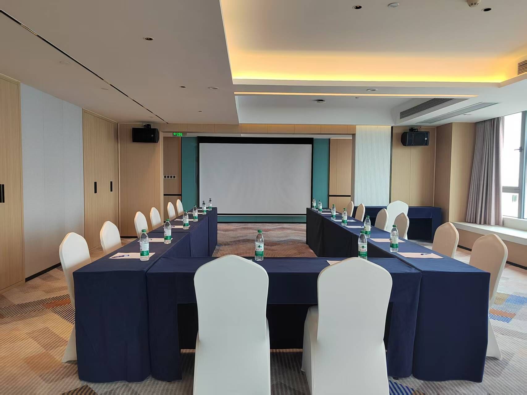 Un salón de conferencias con una larga mesa, sillas blancas y una pantalla para proyector.