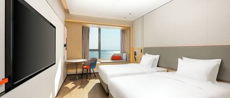 A hotel room with two beds, a desk, a chair, a TV, and a window with a view of the sea.