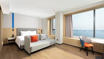 A modern hotel room with a bed, a desk, and a view of the sea.