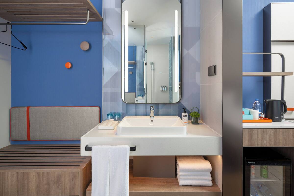A modern bathroom with a large mirror, sink, and shower. There is a towel rack, a small plant, and a coffee maker on the counter.