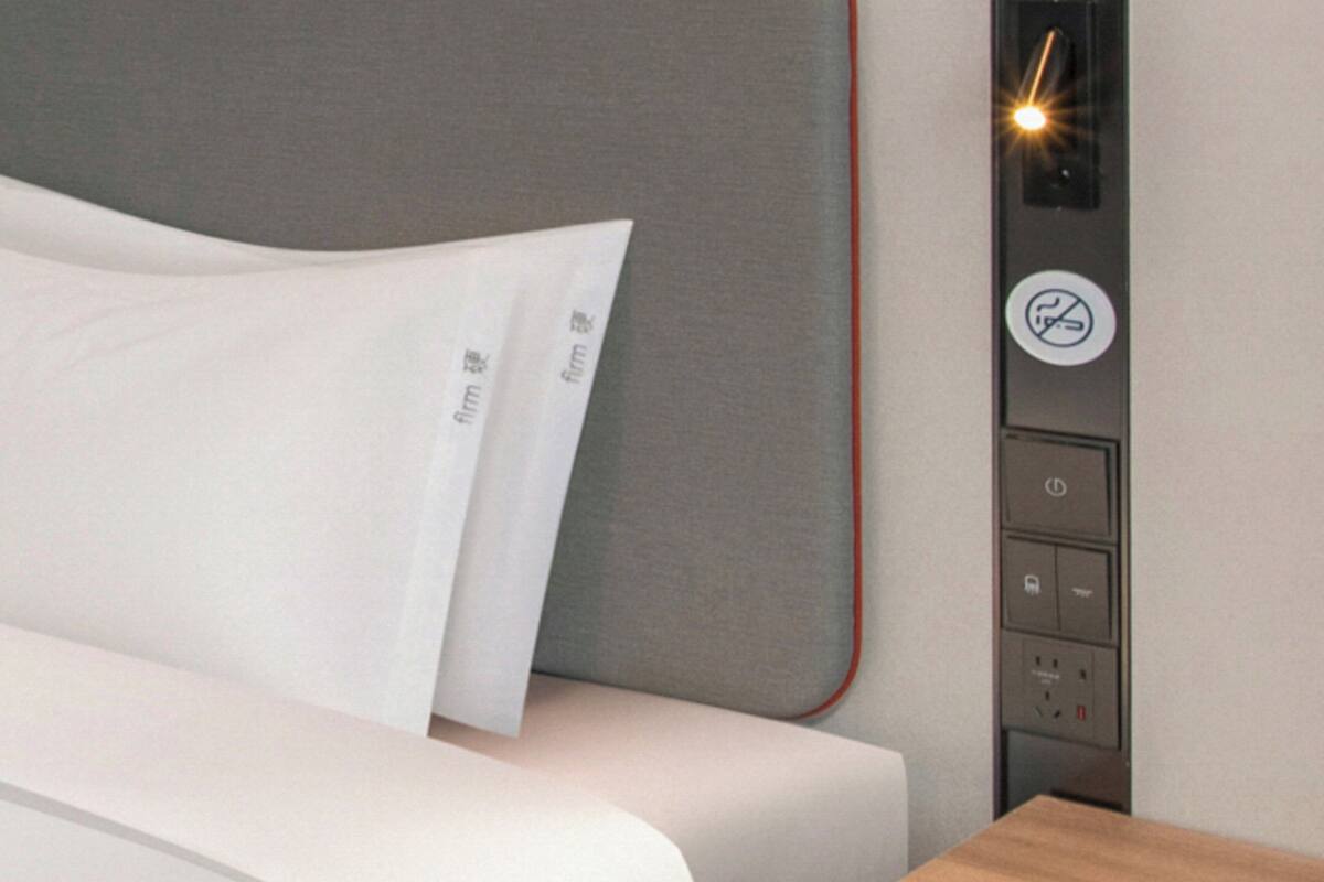 A bed with a white pillow and a grey headboard. There is a control panel with a no-smoking sign and buttons.