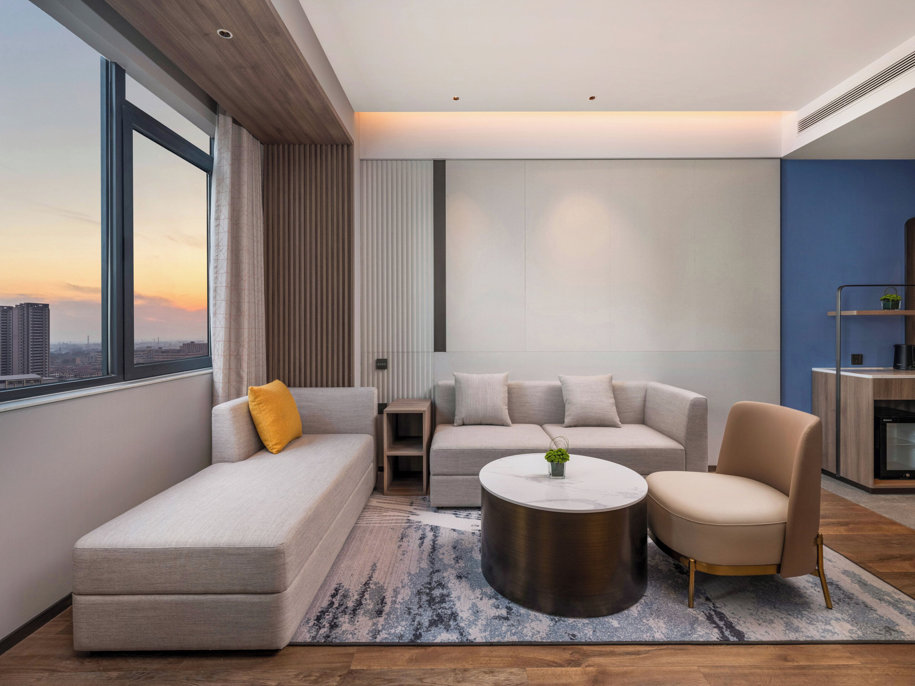 Un salón moderno con sofá, sillón y mesa de centro, vista panorámica de la ciudad a través de ventanas amplias, y una pared decorada en azul.