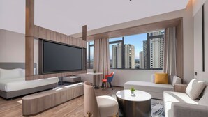 Et moderne hotelværelse med et stort fladskærms-tv, bløde siddepladser, et lille rundt bord og udsigt til høje bygninger.