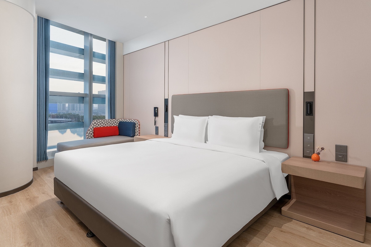 Habitación de hotel moderna con una cama grande, una mesita de noche y vistas a la ciudad a través de amplios ventanales.