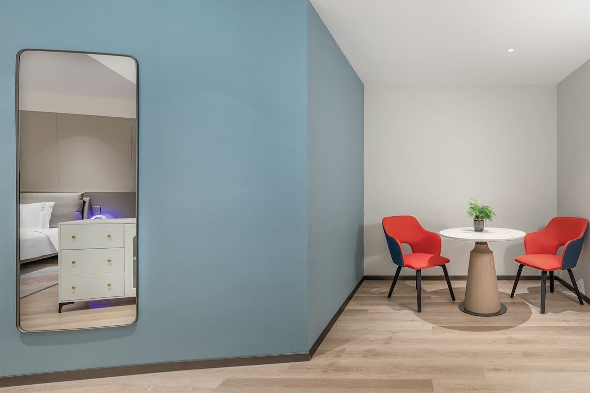 Una habitación moderna con un espejo grande, una cama, una mesita con dos sillas y una planta en maceta.
