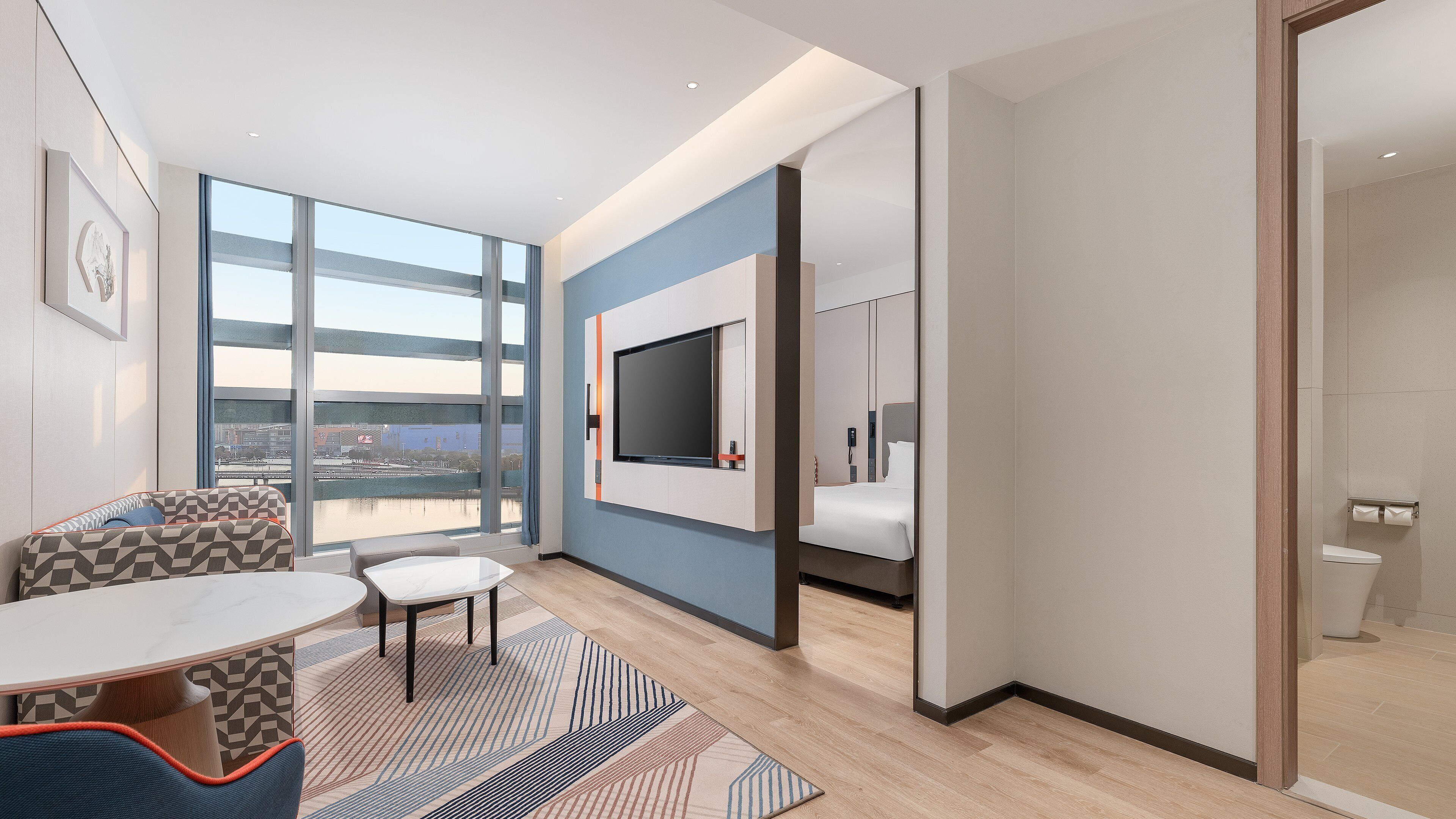 A modern hotel room with a large window, a flat-screen TV, a bed, and a bathroom visible through an open door.