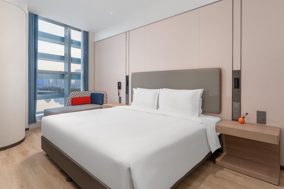 Habitación de hotel moderna con una cama grande, una mesita de noche y vistas a la ciudad a través de amplios ventanales.