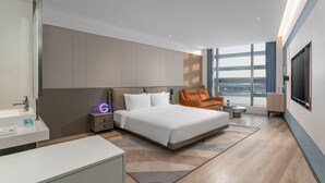 A modern hotel room with a large bed, a sitting area with a chair, a desk, and a large window offering a city view.