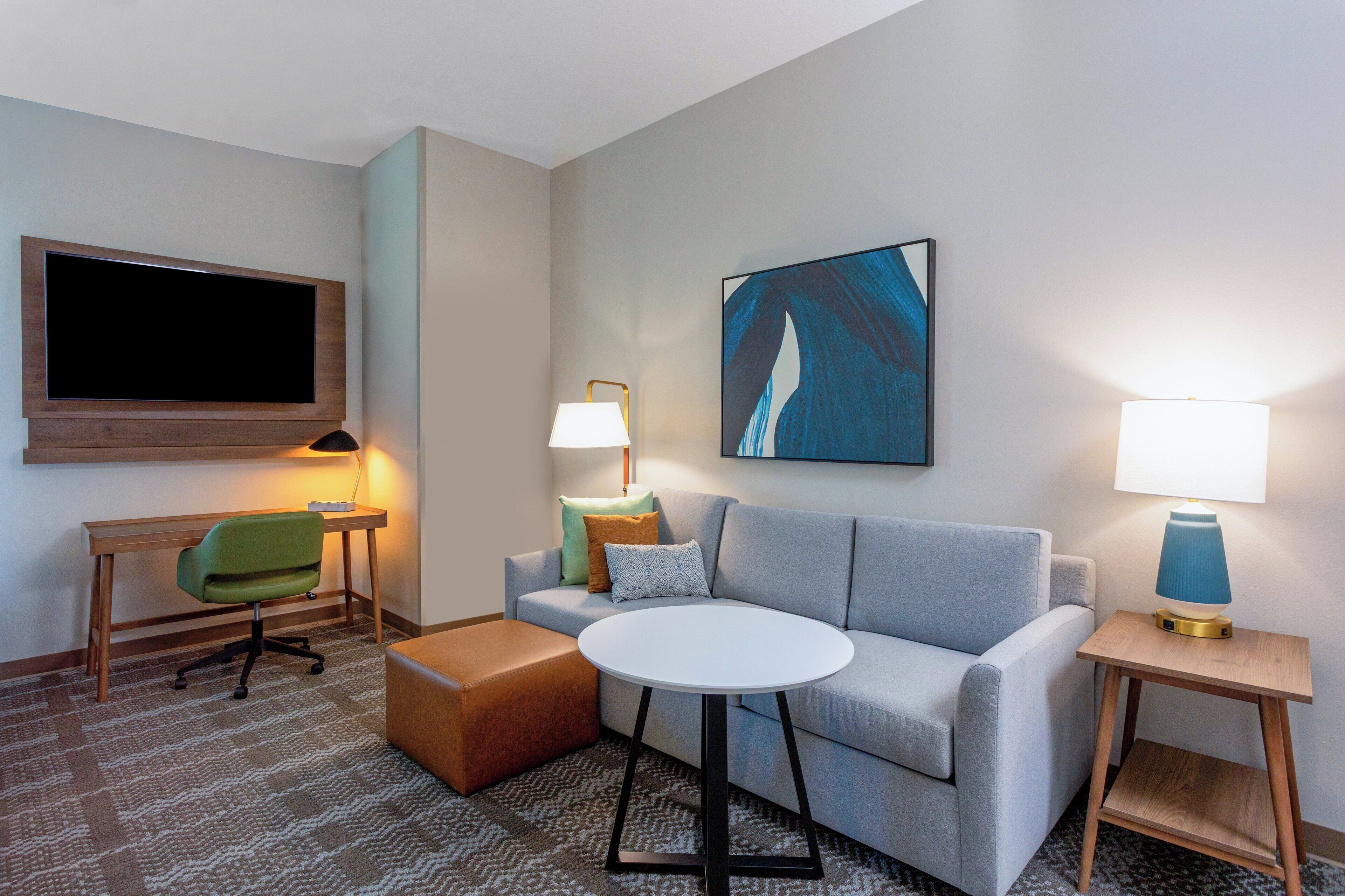 A modern hotel room with a grey sofa, a small round table, a green office chair, a flat-screen TV, two bedside lamps, and a large abstract painting on the wall.