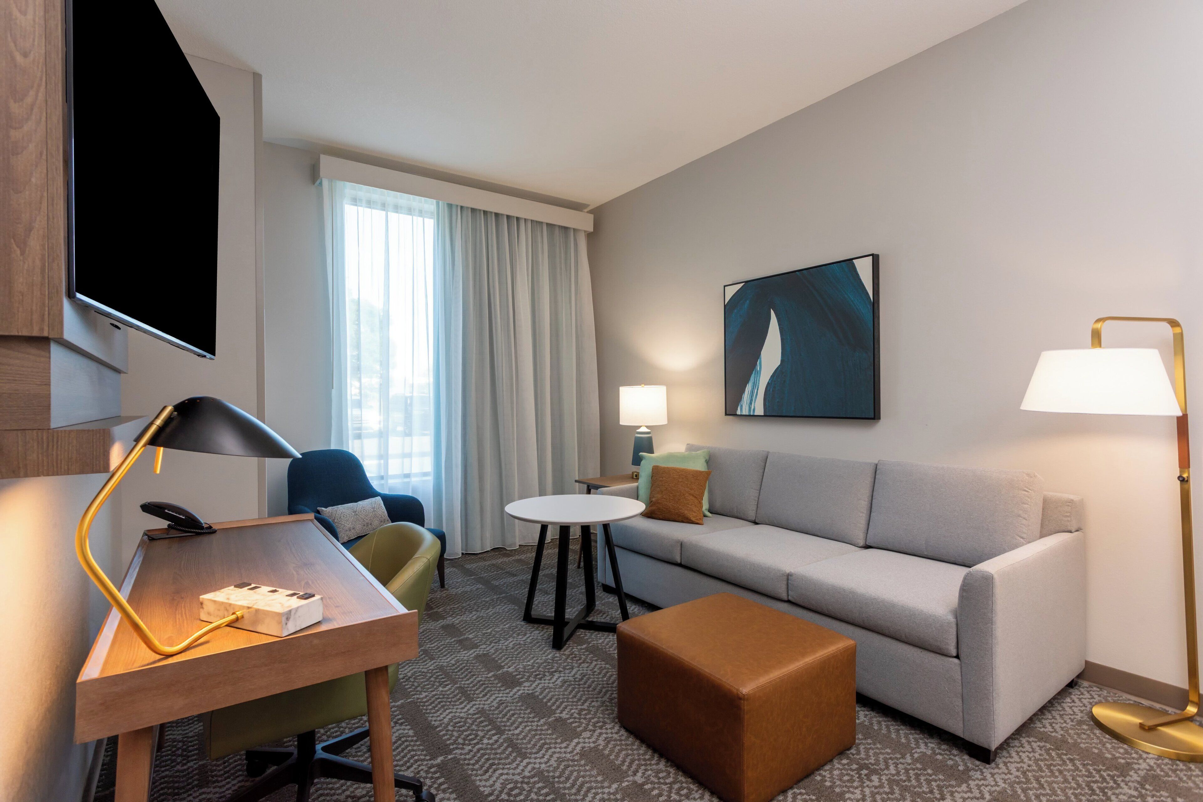 A hotel room with a sofa, a small round table, a desk with a lamp, a television, and a painting on the wall.