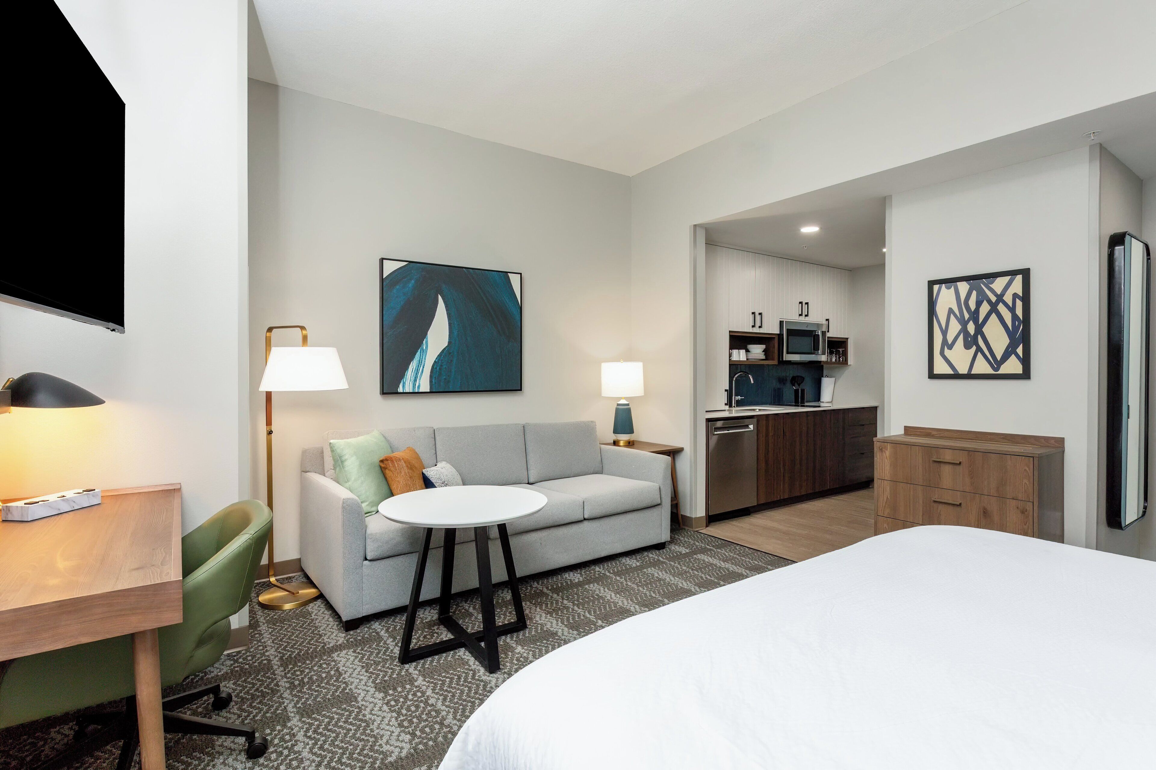 A modern hotel room with a bed, a sofa, a small table, a desk, a lamp, a TV, a kitchen area, and artwork on the walls.