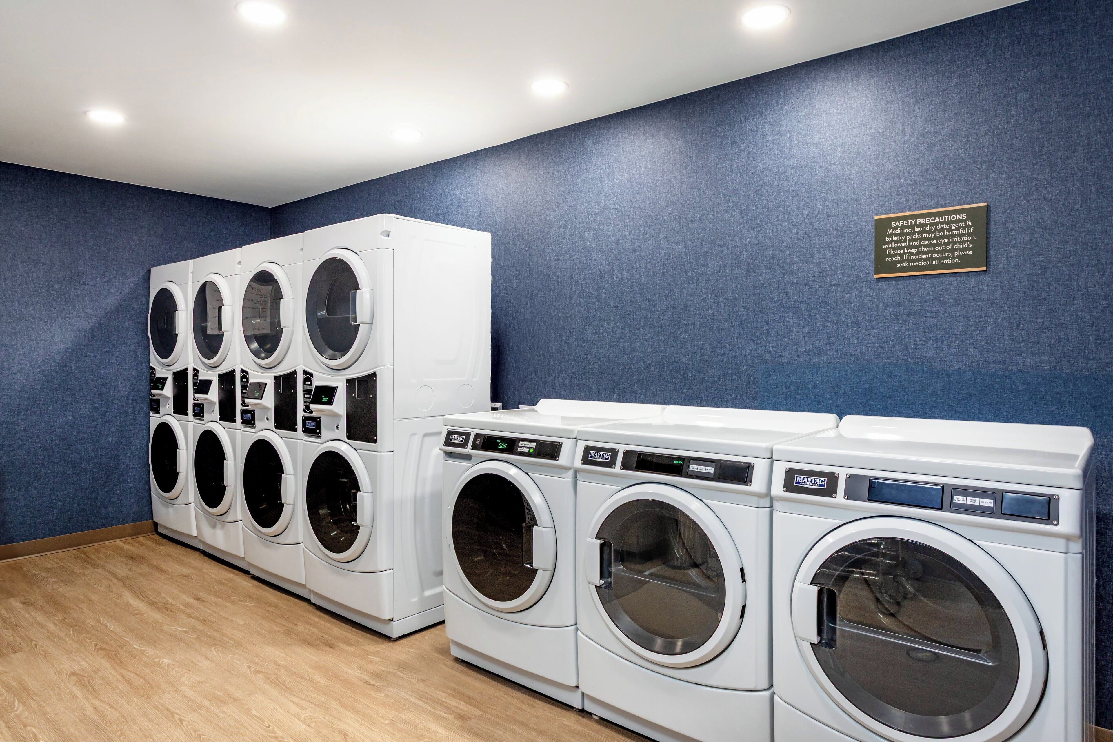 A laundry facility with multiple washing machines and dryers, a safety precautions sign, and a blue wall.