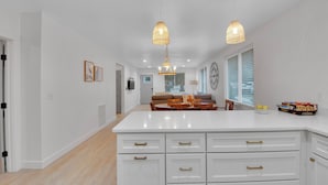 Una cocina moderna con armarios blancos, una isla central, iluminación colgante y una zona de comedor con mesa y sillas.