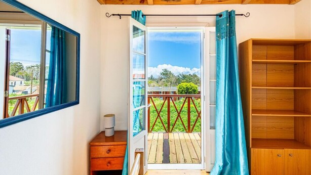Ein Zimmer mit Holzboden, einem Fenster mit blauen Vorhängen, einer Holzregaleinrichtung und Blick auf eine Terrasse und Grünflächen.