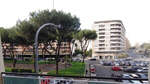 Vue depuis un balcon donnant sur un parking où se trouvent plusieurs voitures, un rond-point et, au loin, un immeuble à plusieurs étages.