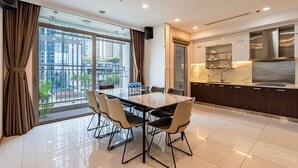 A modern dining area with a marble table and chairs, a kitchen with dark cabinetry and stainless steel appliances, and large windows offering a city view. - 4 Bedroom Apartment for Rent at Vinhomes Central Park (Binh Thanh District)