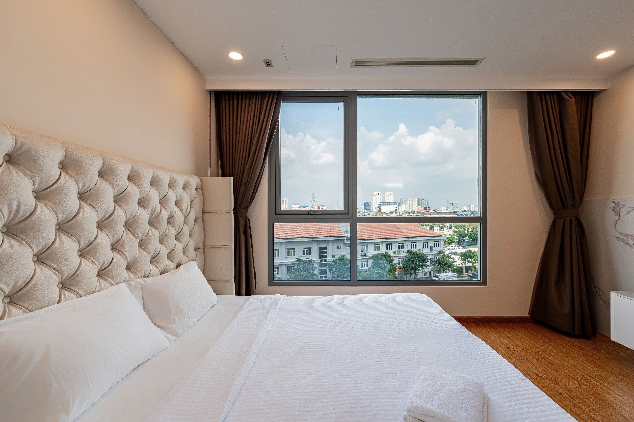 Una habitación de hotel moderna con una cama grande, un cabecero acolchado y vistas a la ciudad a través de amplios ventanales.