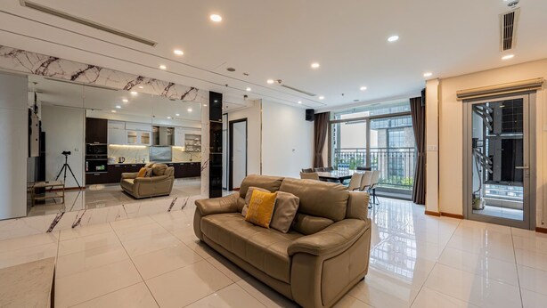 A spacious living area with a large sofa, a dining table, and a kitchen area visible through a glass partition. - 4 Bedroom Apartment for Rent at Vinhomes Central Park (Binh Thanh District)