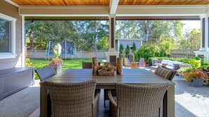 Un patio al aire libre con techo de madera, una mesa de comedor con sillas de mimbre y vista a un jardín trasero con cercado y follaje.