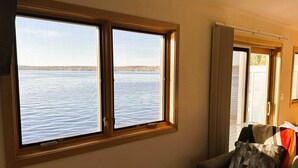 A room with a large window overlooking a body of water, a chair with cushions, and a door with curtains. - Lakefront Views + Walk to Dining | Kayaks + Firepit (Pewaukee)