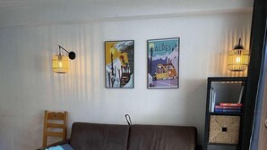 Een woonkamer met een bruine bank, een houten salontafel en twee wandlampen. Daarboven hangen twee kunstwerken en rechts bevindt zich een boekenkast met boeken en decoratieve objecten.