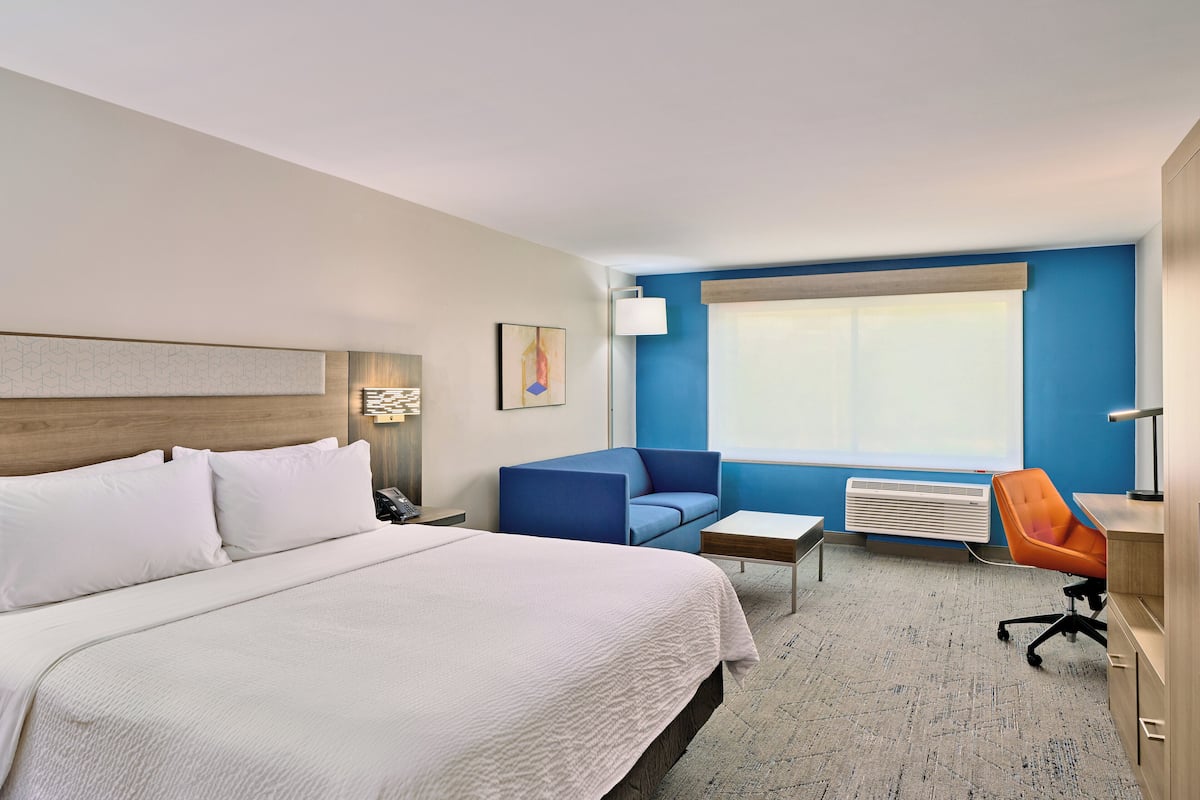 A hotel room with a large bed, a blue sofa, an orange office chair, a small table, and a window with blinds.