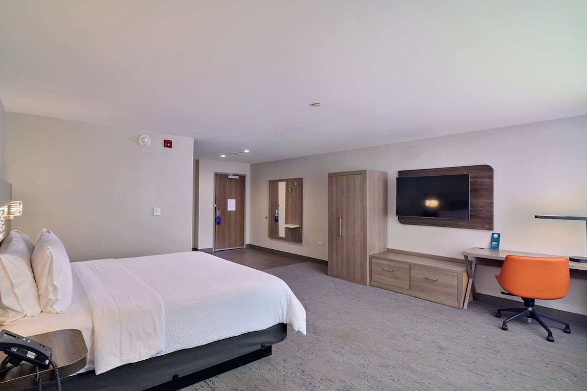 A hotel room with a bed, a desk, an orange office chair, a TV mounted on the wall, and a wardrobe.