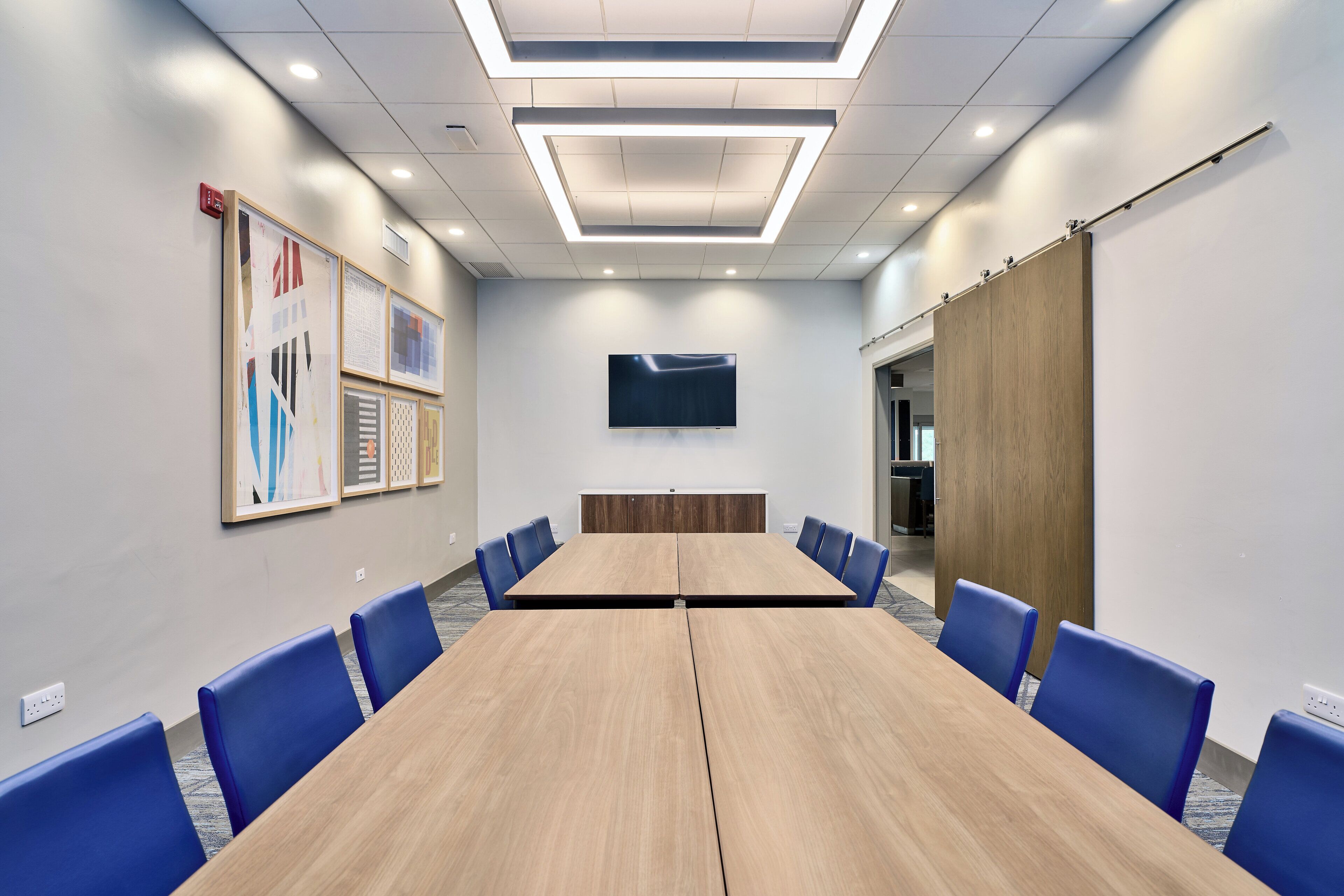 A conference room with a long table, blue chairs, a flat-screen TV, and framed artwork on the walls.