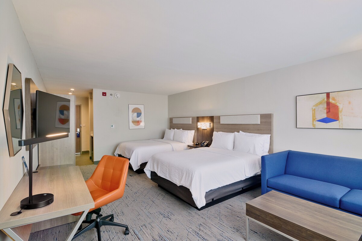 A hotel room with two beds, a desk with a lamp, an orange chair, a blue sofa, and a TV.