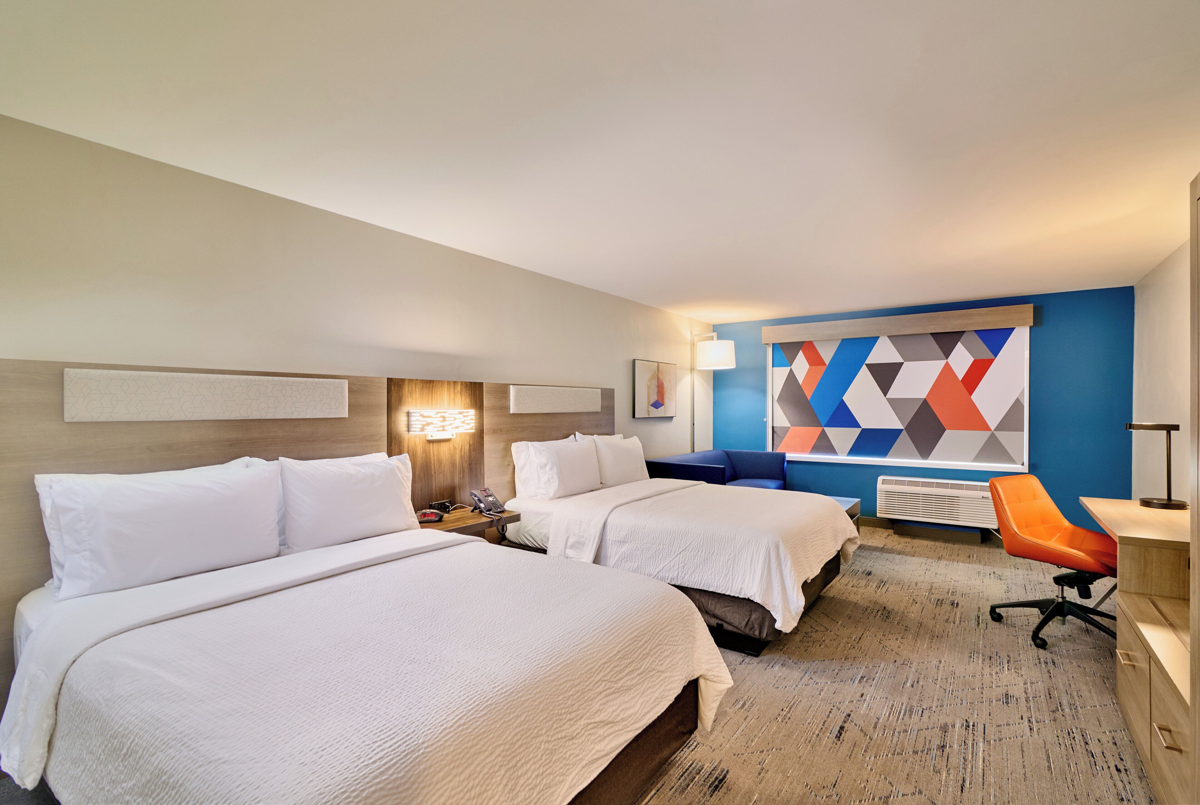 A hotel room with two beds, a desk, a chair, a lamp, and a wall with a geometric pattern.