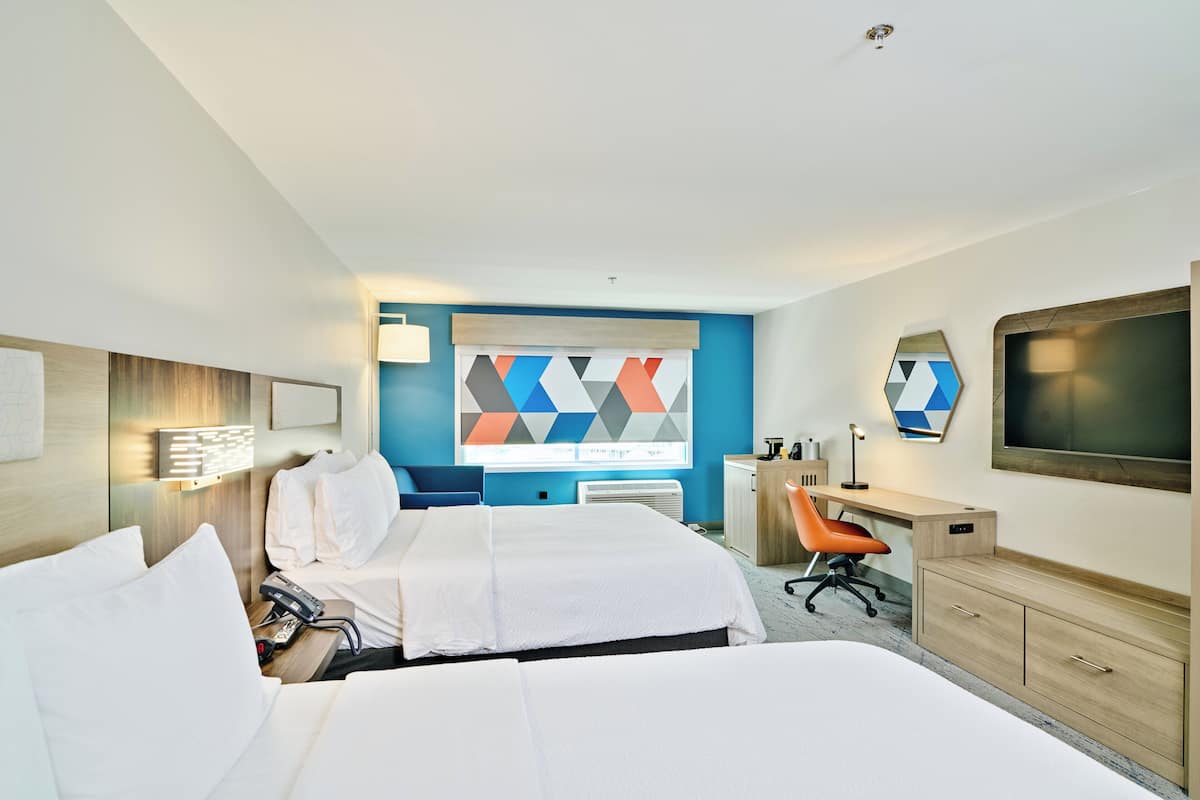 A hotel room with two beds, a desk with a chair, a TV, and a large geometric artwork on the wall.