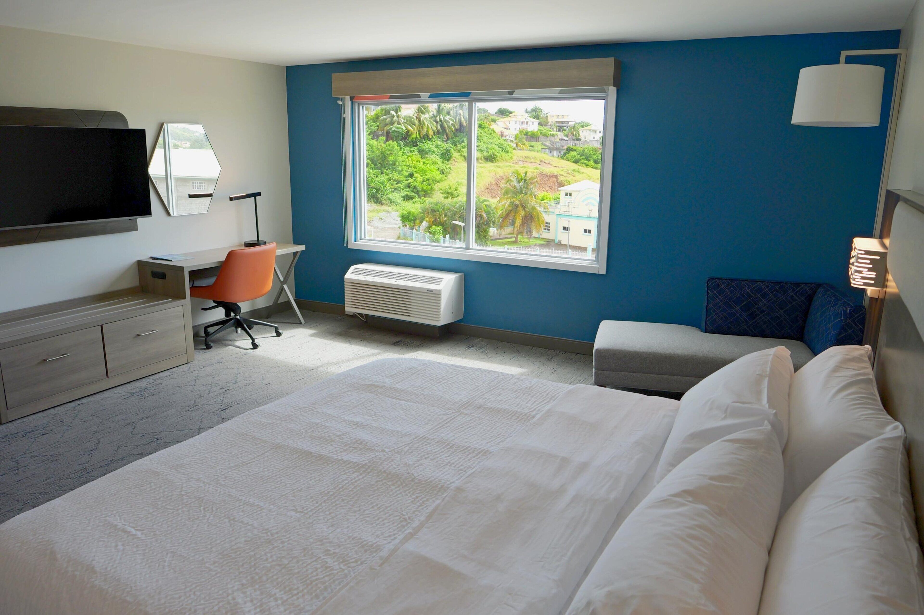 Habitación de hotel con cama, escritorio, silla, televisor y ventana que ofrece vistas a zonas verdes y edificios.
