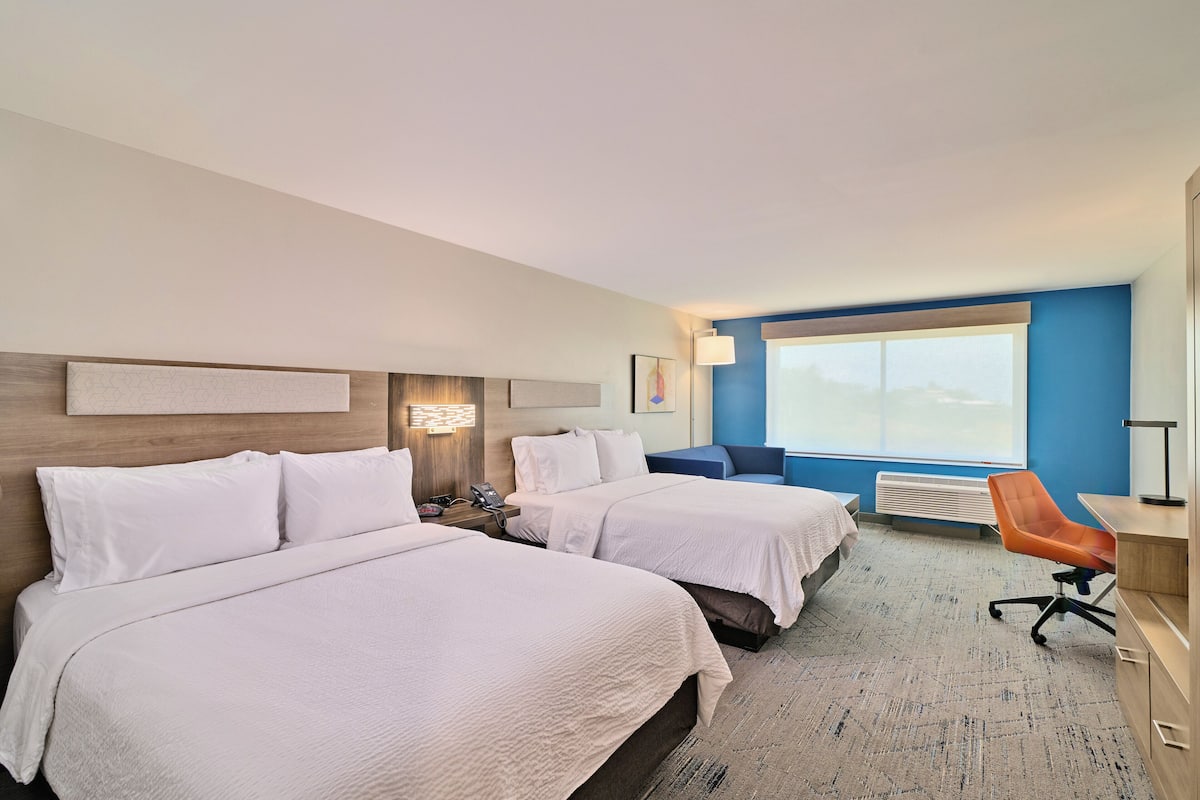 A hotel room with two beds, a desk, an orange chair, a large window, and a wall-mounted heater.