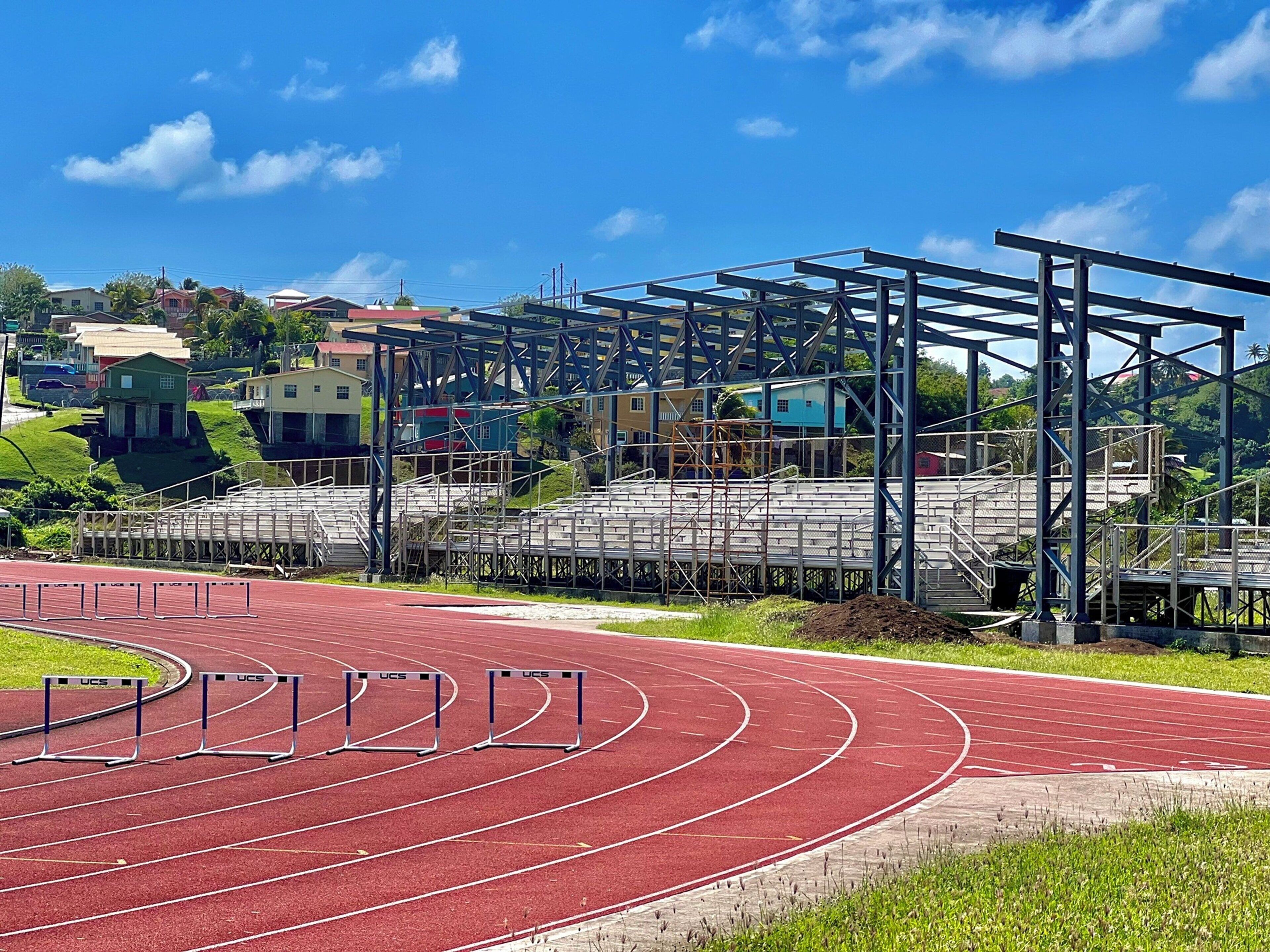 Pista de atletismo con vallas, una estructura metálica azul y casas al fondo.
