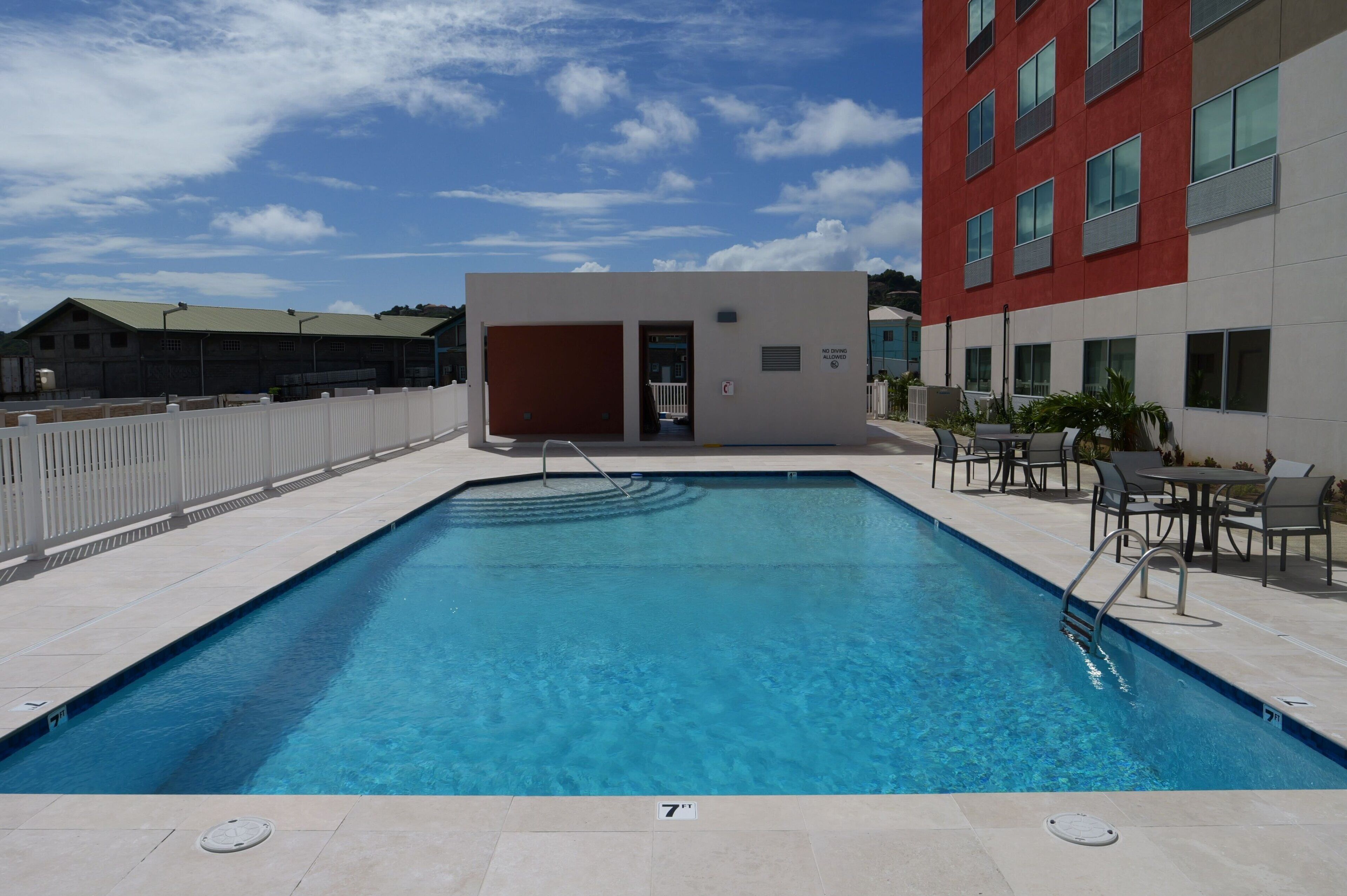 Una piscina rettangolare con indicatore di profondità di 18 centimetri, circondata da una recinzione bianca, con un edificio in lontananza caratterizzato da una facciata rossa e numerose finestre.