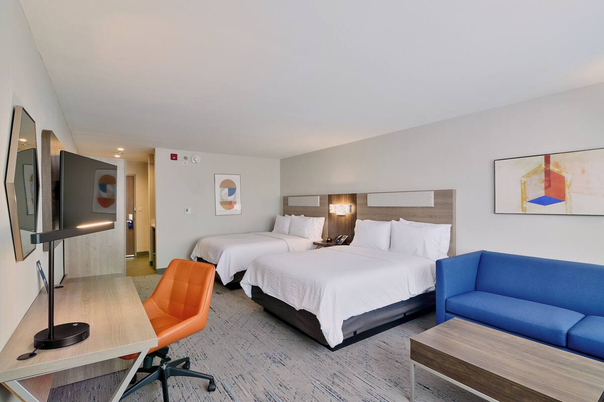 A hotel room with two beds, a desk with a lamp, an orange chair, a blue sofa, and a TV.