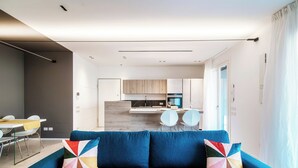 Ein modernes Wohnzimmer mit einem blauen Sofa und einem farbenfrohen Kissen, ein Essbereich mit Tisch und Stühlen sowie eine Küche im Hintergrund.