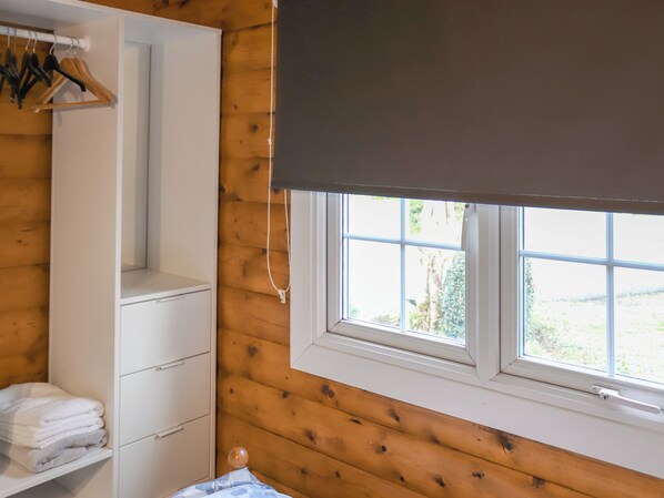 Una habitación con paneles de madera, un armario blanco, una ventana con persiana blanca y vista a zonas verdes.