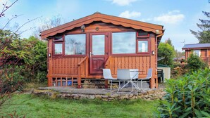 A wooden cabin with a stone foundation, a porch with chairs, and a small table. - Babbling Brook Lodge (Trawsfynydd)