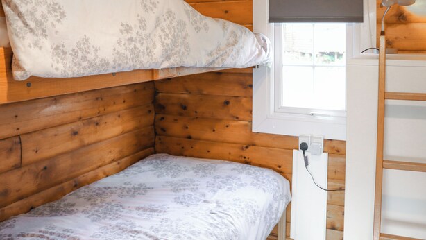 Une pièce dans un chalet en bois, avec un lit superposé, une fenêtre avec un store et un bureau avec une lampe.