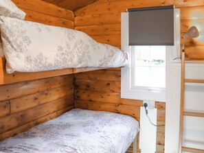 A wooden cabin room with a bunk bed, a window with a blind, and a desk with a lamp.