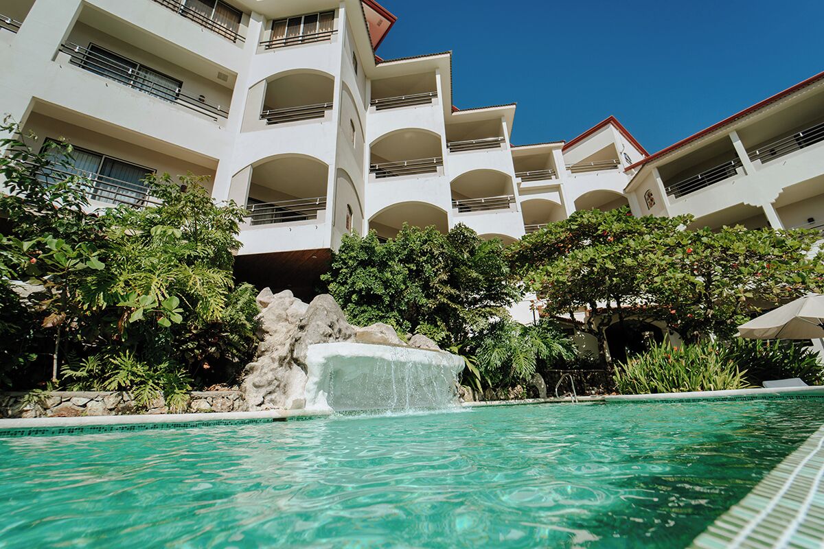 A hotel with a swimming pool, surrounded by lush greenery and a rock waterfall feature.