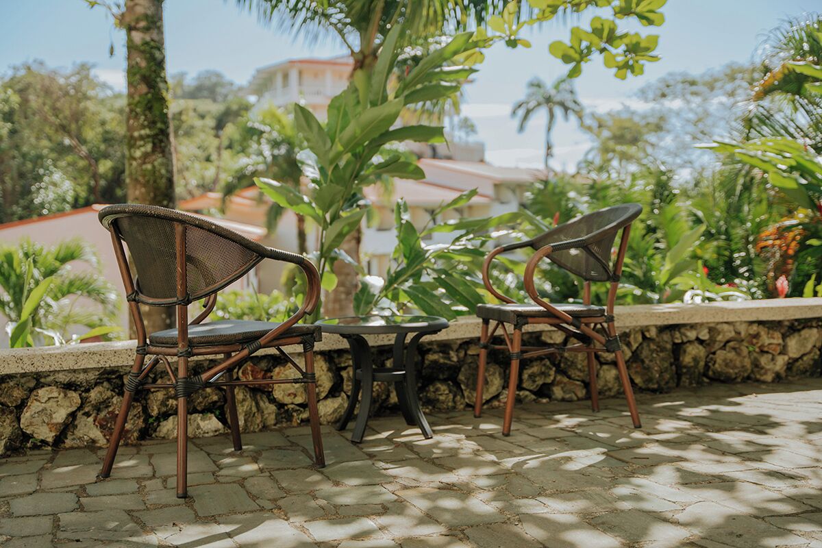 A patio area with wicker chairs and a table, surrounded by greenery and a stone wall.