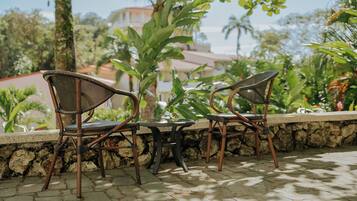 A patio area with wicker chairs and a table, surrounded by greenery and a stone wall.