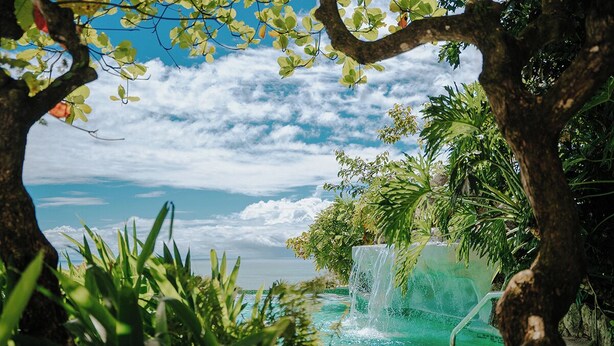 A pool with a waterfall, surrounded by lush greenery and a view of the ocean. - Vista Suites at Parador (Quepos)
