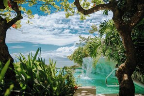 A pool with a waterfall, surrounded by lush greenery and a view of the ocean.