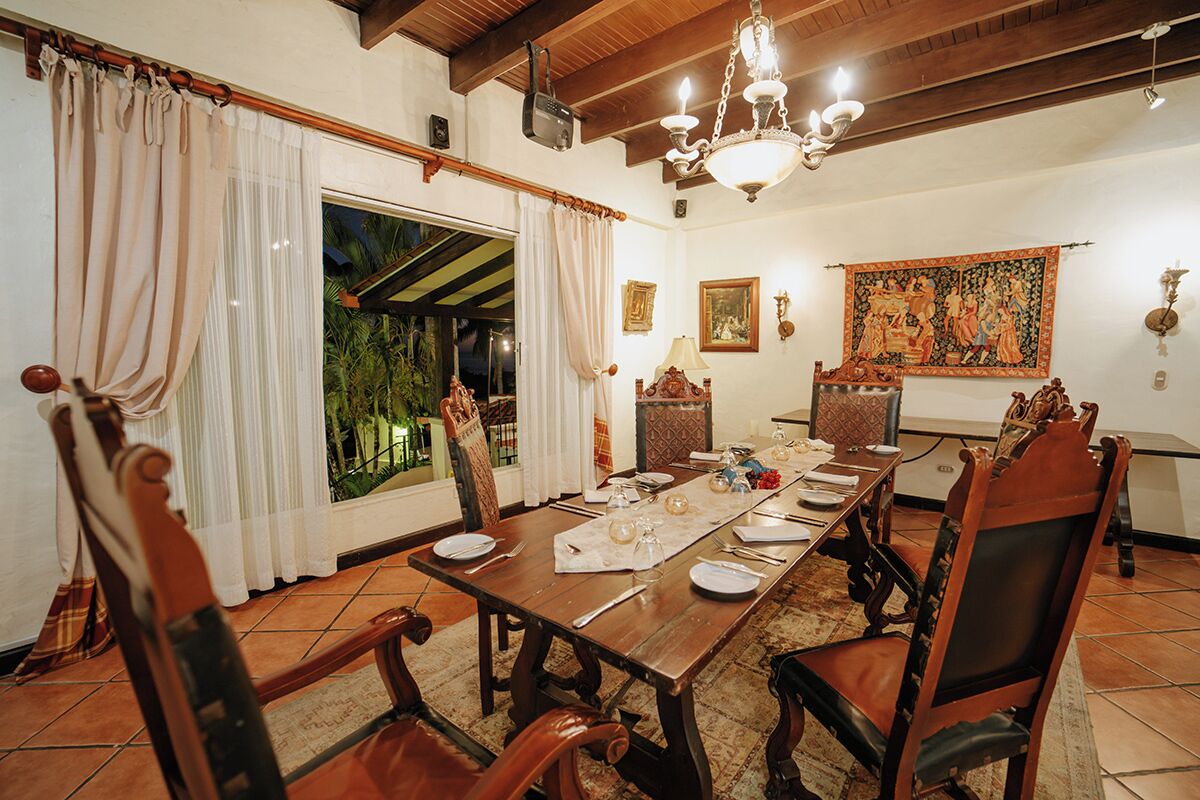 A dining room with a wooden table set for six, surrounded by ornate chairs, a chandelier, and a large tapestry on the wall.